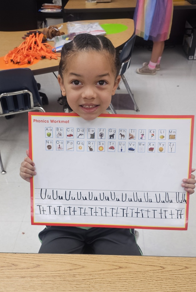 A young child displays a worksheet with alphabets, numbers, and pictures in a classroom setting.