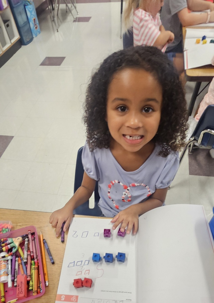 A child sits at a table, smiling, and holds a book with a counting exercise. Behind them, other children work at desks.