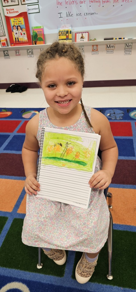 A child in a white dress sits and smiles, holding a drawing of a tree. Background includes a classroom with a whiteboard.