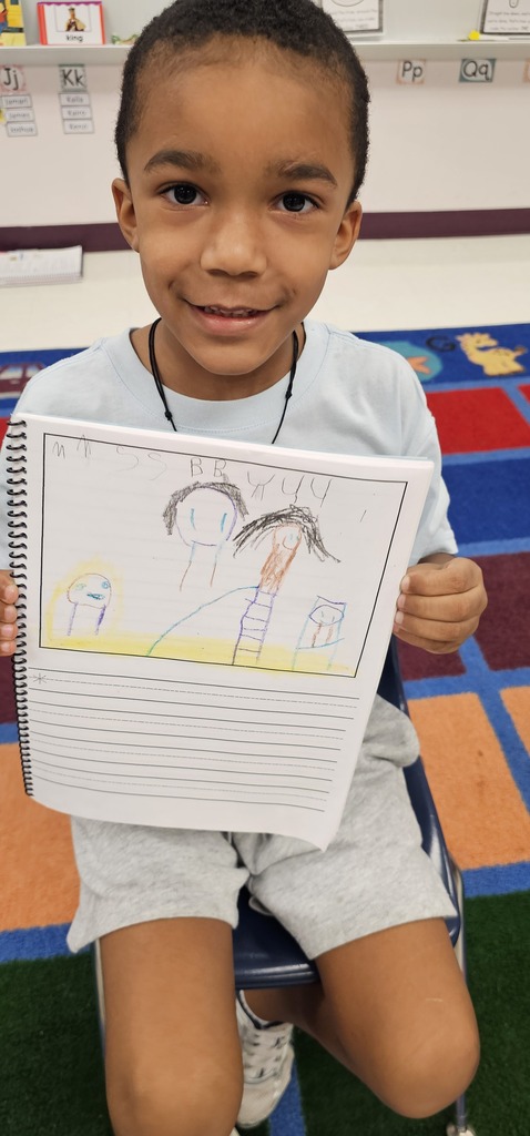 A boy in a classroom holds up a drawing. He is seated on a chair. The floor is multicolored.