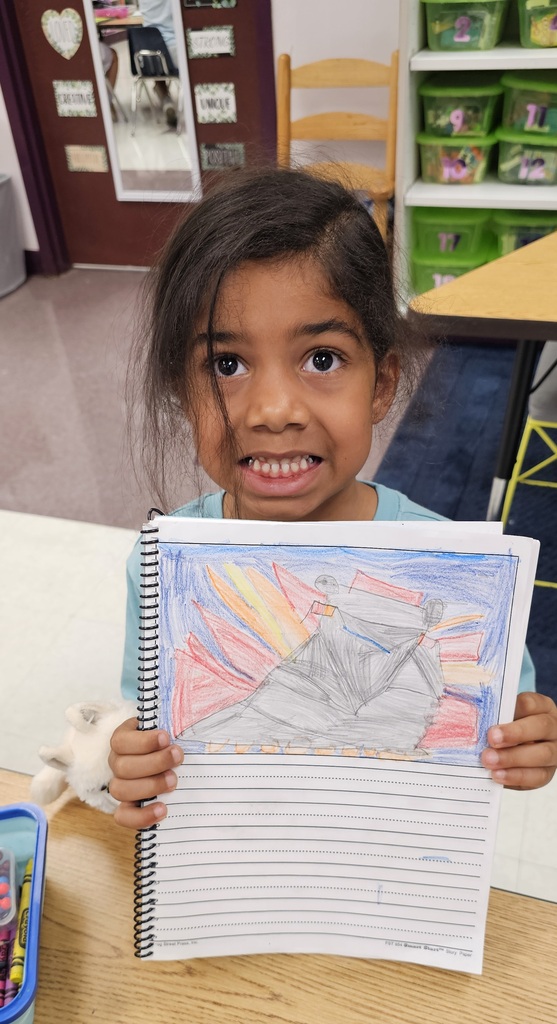 A young child holds a colorful drawing of a mountain and boat in a classroom. Behind them, shelves with storage bins and a wooden chair are visible.