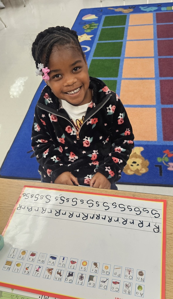 A young girl in a floral-printed jacket sits at a table with a colorful rug behind her. She holds a paper with alphabet letters and pictures.