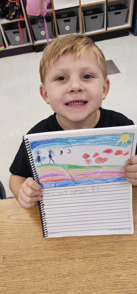 A child in a black shirt holds up a notebook with a drawing of a landscape.