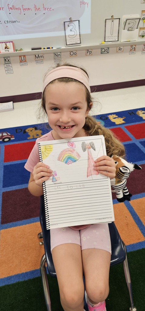 A child holding a colorful drawing with a rainbow, seated in a classroom with a carpeted floor.