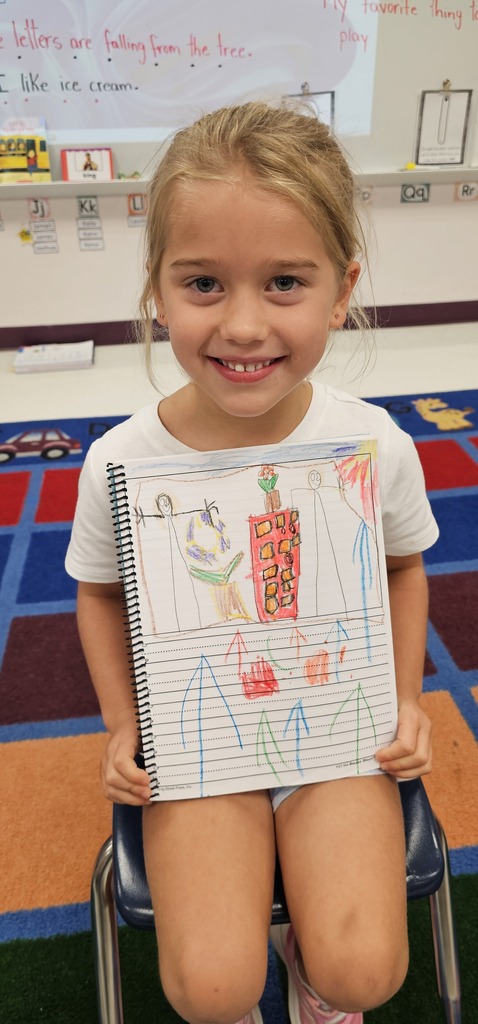 A girl holding a notebook with drawings and smiling, seated on a chair in a classroom.