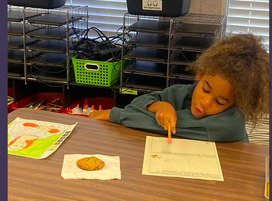 Child with curly hair at a desk, holding a cookie and coloring a worksheet. Background includes storage shelves and blinds.