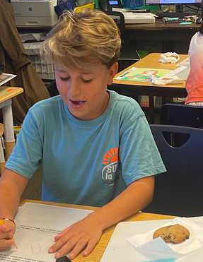 A boy in a light blue shirt focuses on a worksheet, holding a pencil. Nearby, a cookie rests on a napkin.