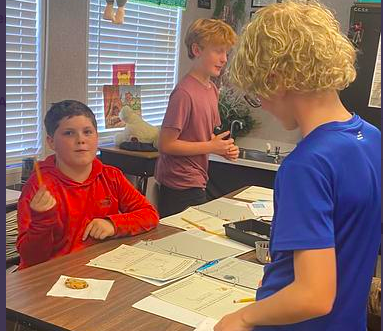Three boys in a classroom setting, two standing at a table, one holding a pencil. Papers, cookies, and a stuffed animal visible.