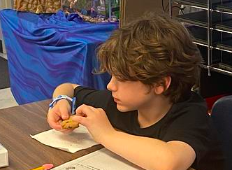 A young child with short hair, wearing a black shirt, sits at a desk, examining a cookie.