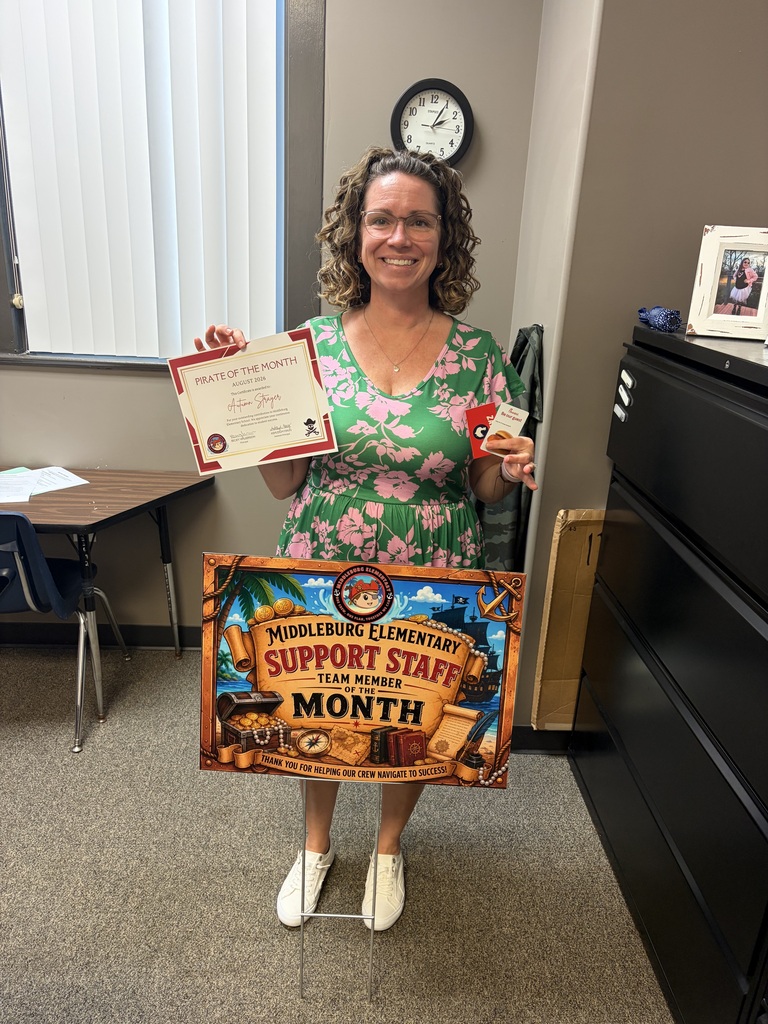 A woman in a floral dress and glasses holds a certificate and a sign that says Support Staff of the Month. She stands in an office with a desk and a clock.