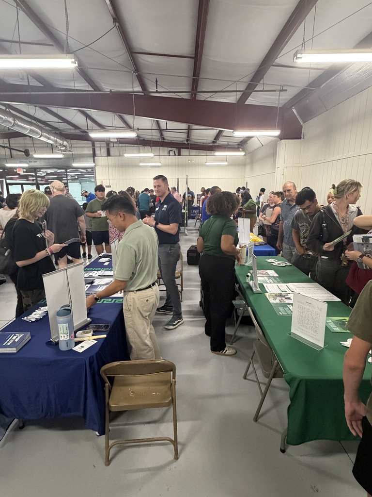 People gather at tables in a room, some standing and some seated. Tables have papers and folders.