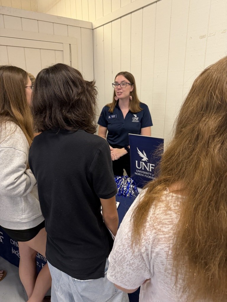 Five people standing near a table. The person in the center wears glasses and a blue shirt with "UNF" printed on it.