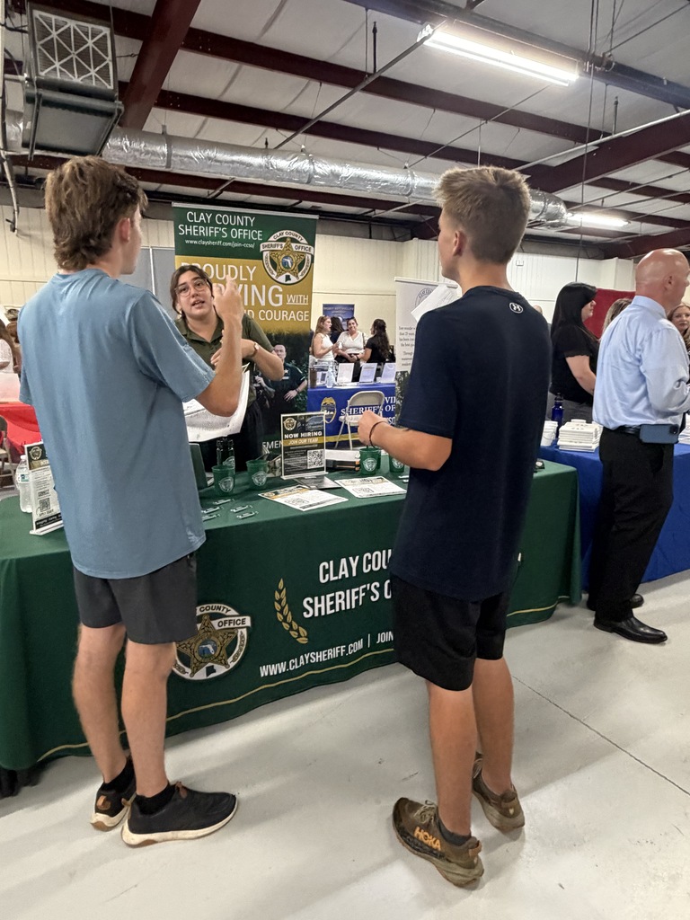 Two boys are standing at a table with a green tablecloth, looking at a woman. Behind them, people are at tables.
