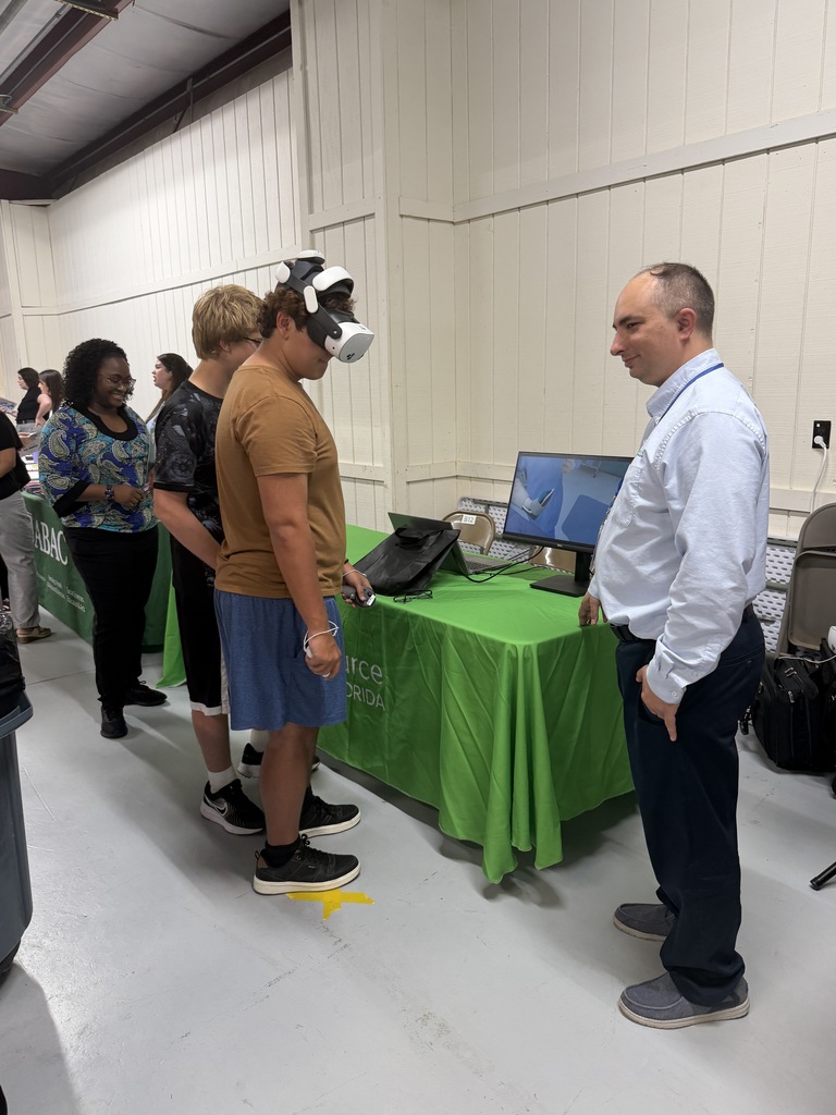 A man wearing a VR headset stands at a table with a monitor, while another man in front of him observes.