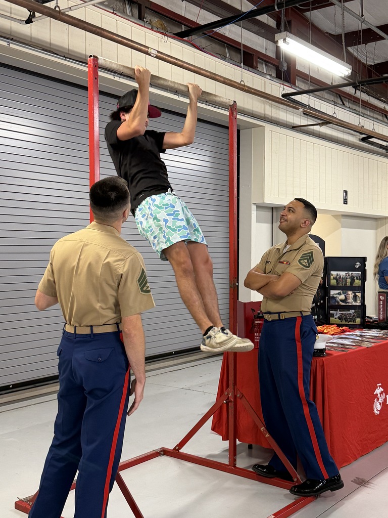 Three men in uniforms. One man is doing pull-ups on a red bar. Two other men watch.