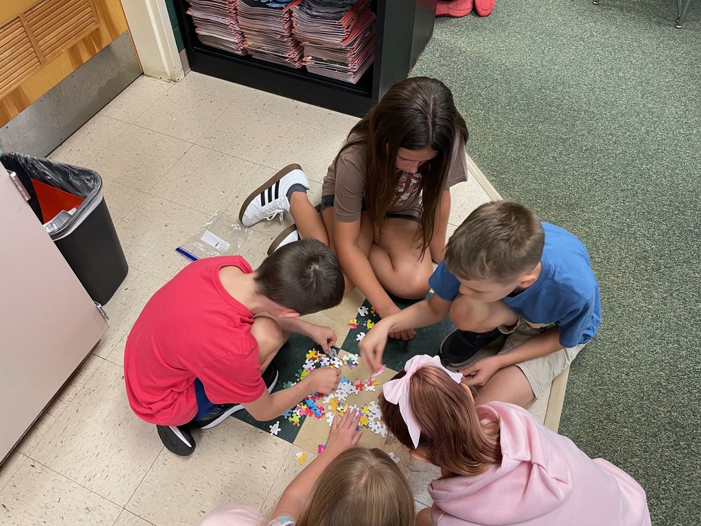 Five children work on a puzzle on a tiled floor. One child wears a headband, and a trash can is nearby.