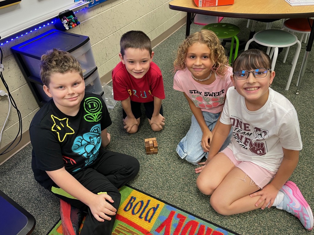 Four children sit on a rug with a wooden block between them. They are in a classroom with a table and chairs.