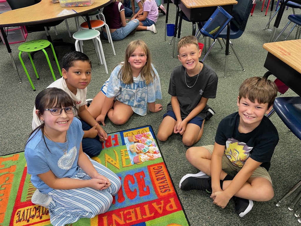 Five children are sitting on a colorful rug, smiling at the camera. A table and chairs are in the background.