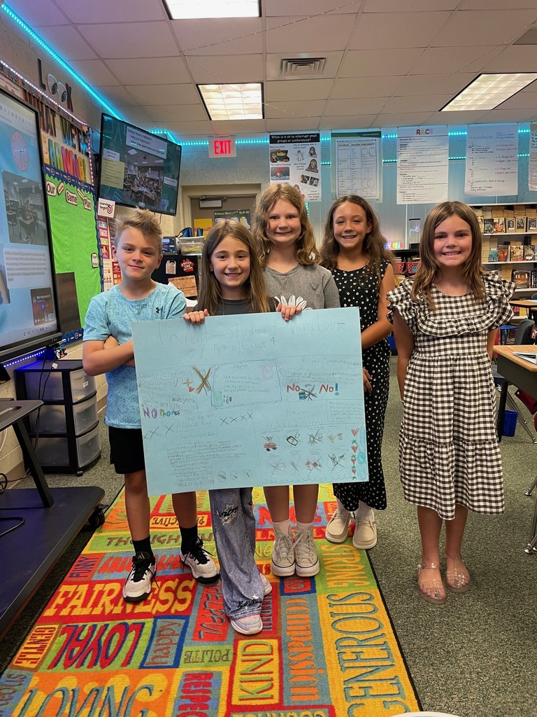 Five children stand in a classroom holding a large blue sign with white letters. Bright colors cover the floor.