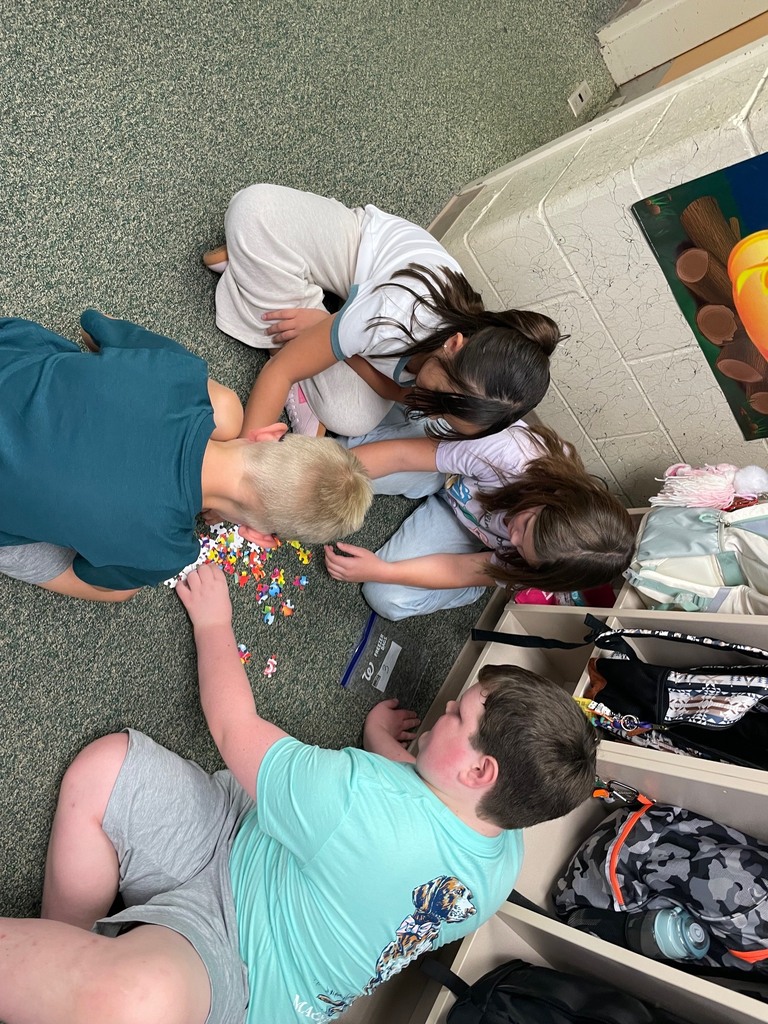 Four people on a floor, engaged in an activity. One uses tweezers, others handle items. A white wall, shelves with objects.