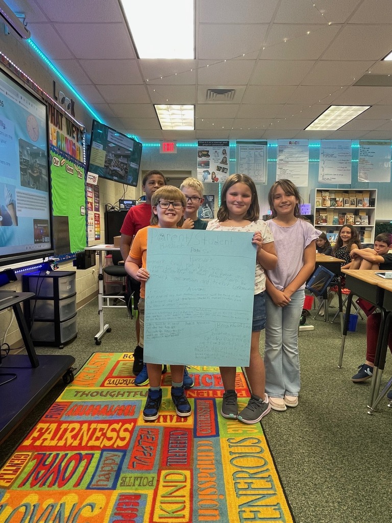 Four children holding a large blue sign with words in a classroom with carpeted floors and desks.
