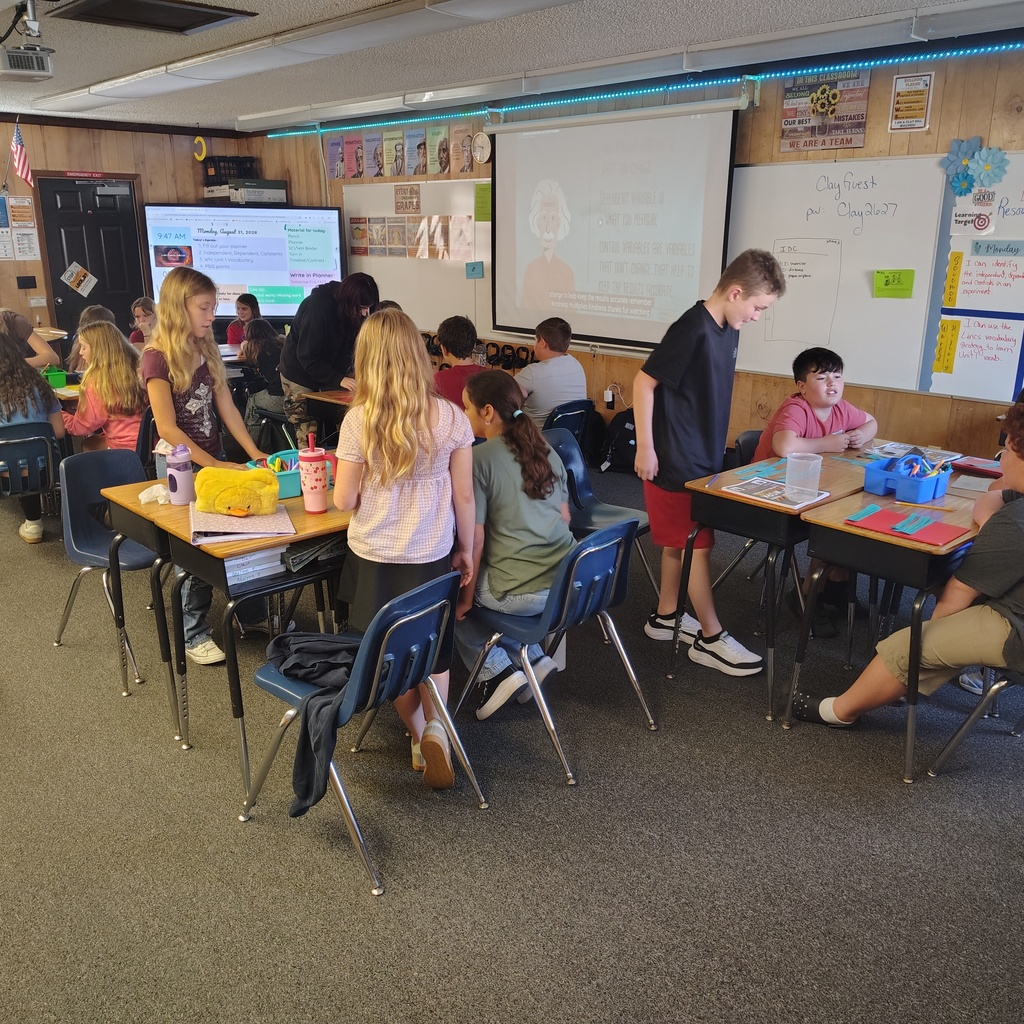 Kids are seated at desks in a classroom. A teacher stands in front of a projector screen.
