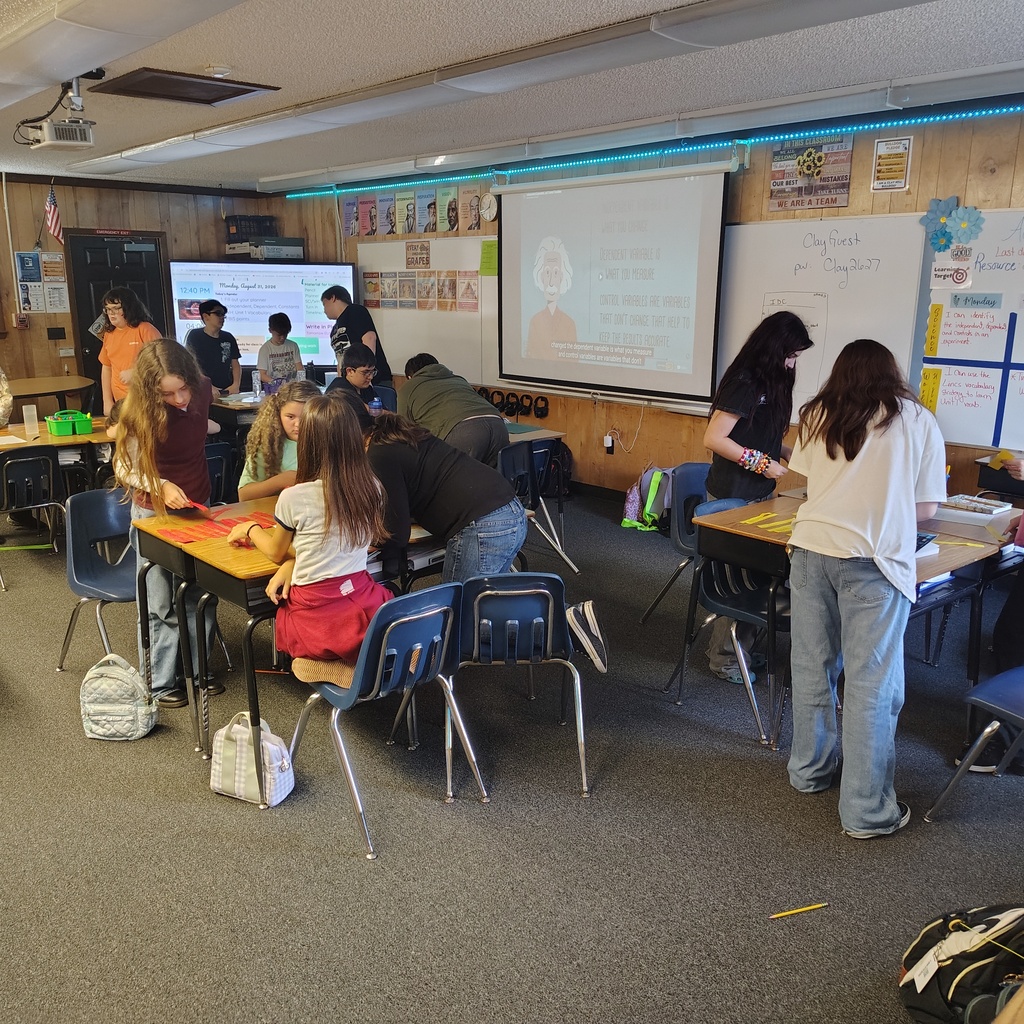 Students seated at tables in a classroom, working on a project. One person is standing at a whiteboard.