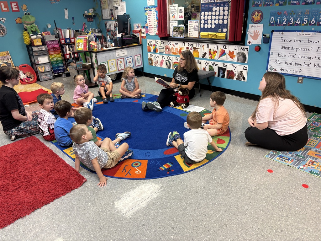 A classroom with blue walls, a woman reading to young children sitting in a circle, and a whiteboard.