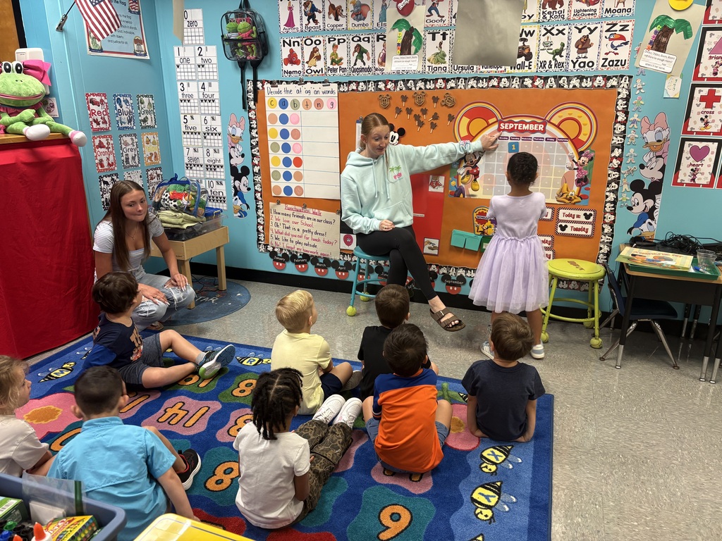 An instructor gestures toward a chart in a classroom with young children sitting on a rug.