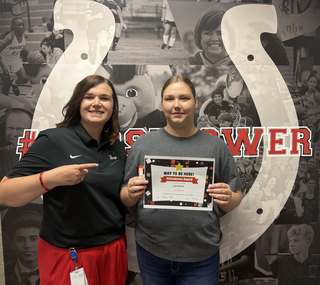 Two women stand in front of a horseshoe mural, one holding a framed certificate, smiling.