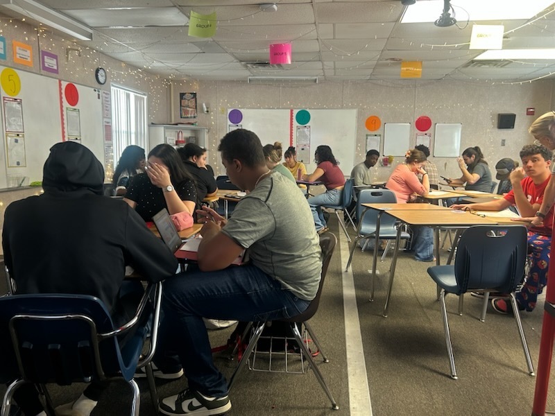 Students sit at tables in a classroom with white boards, colorful balloons, and a white ceiling.