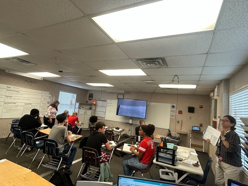 A classroom with several students seated around desks. A teacher stands in front of them. There are whiteboards and monitors mounted on the walls.