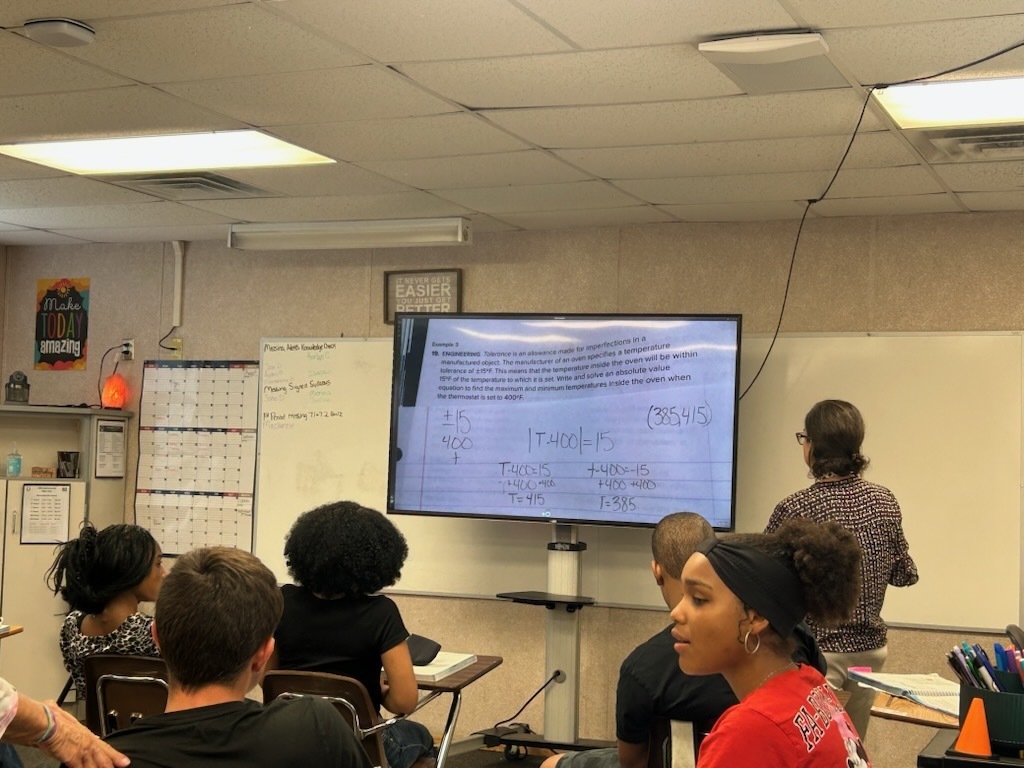 A classroom with students seated. A teacher stands in front of a large screen showing text. The room has whiteboards and a bulletin board.