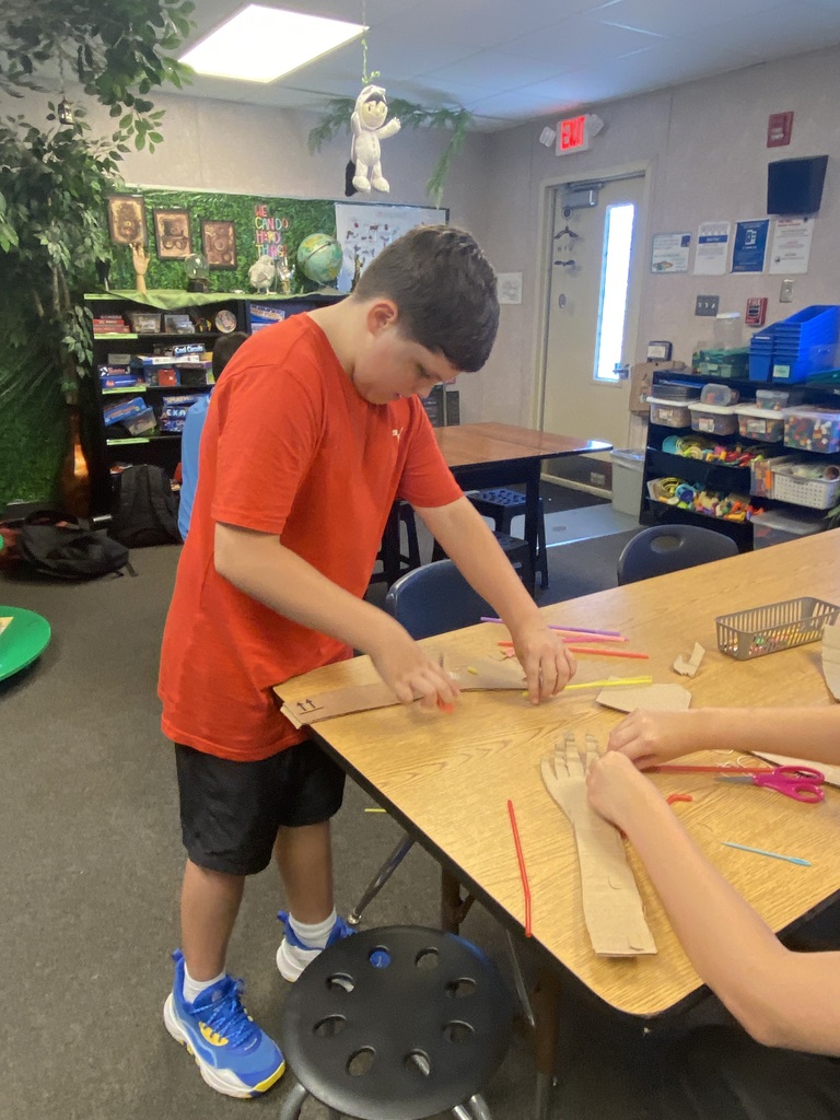A young boy in a red shirt and black shorts works on a craft project at a table.