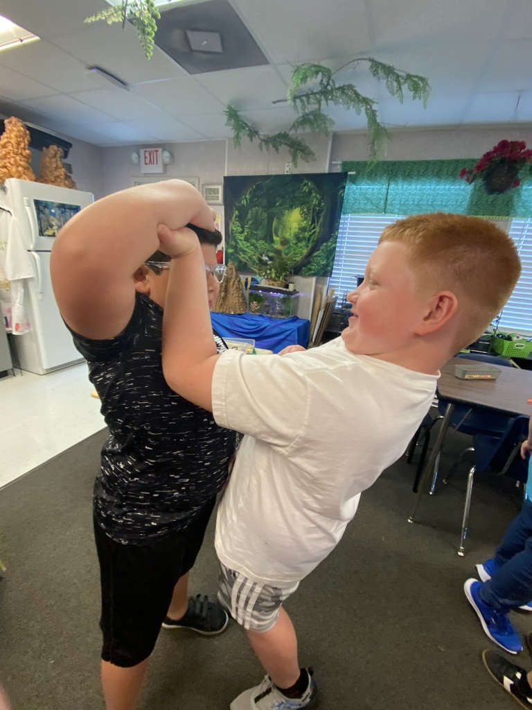 Two boys playfully wrestle in a classroom. One holds a toy sword. A table and chairs are nearby.