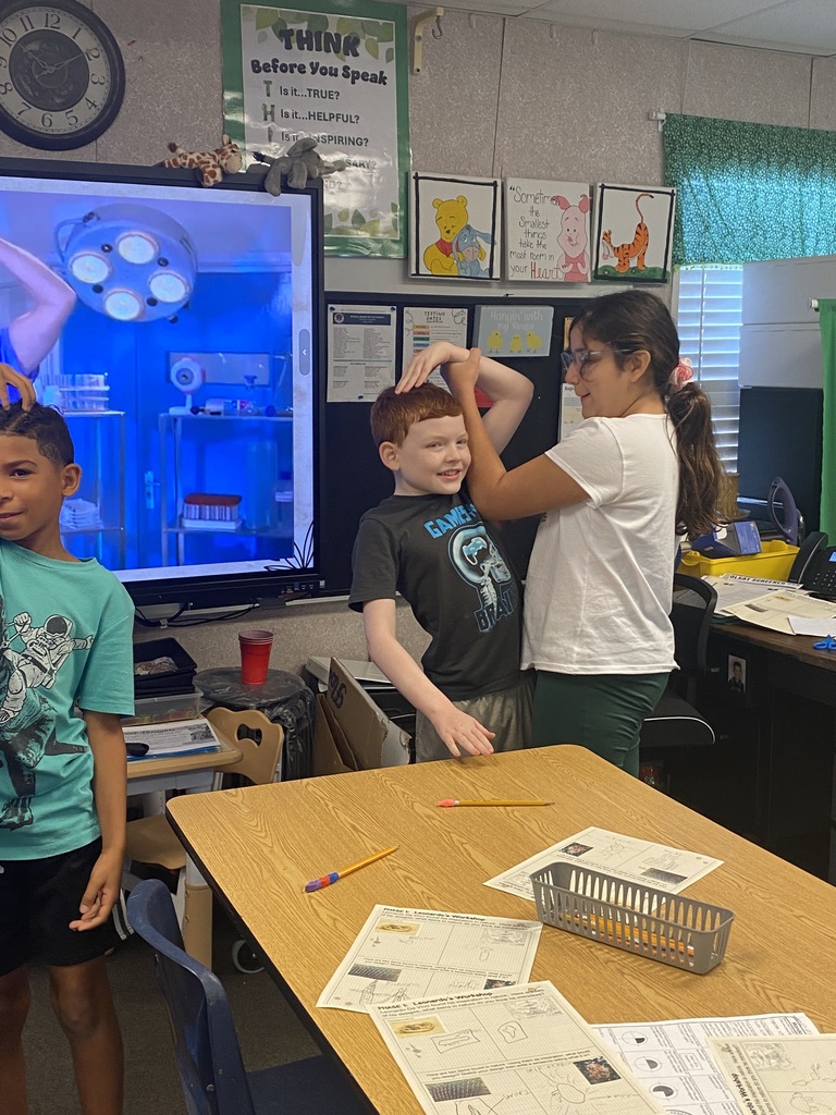 Children in a classroom with a monitor on the wall, one holding a boy's head while another looks on.