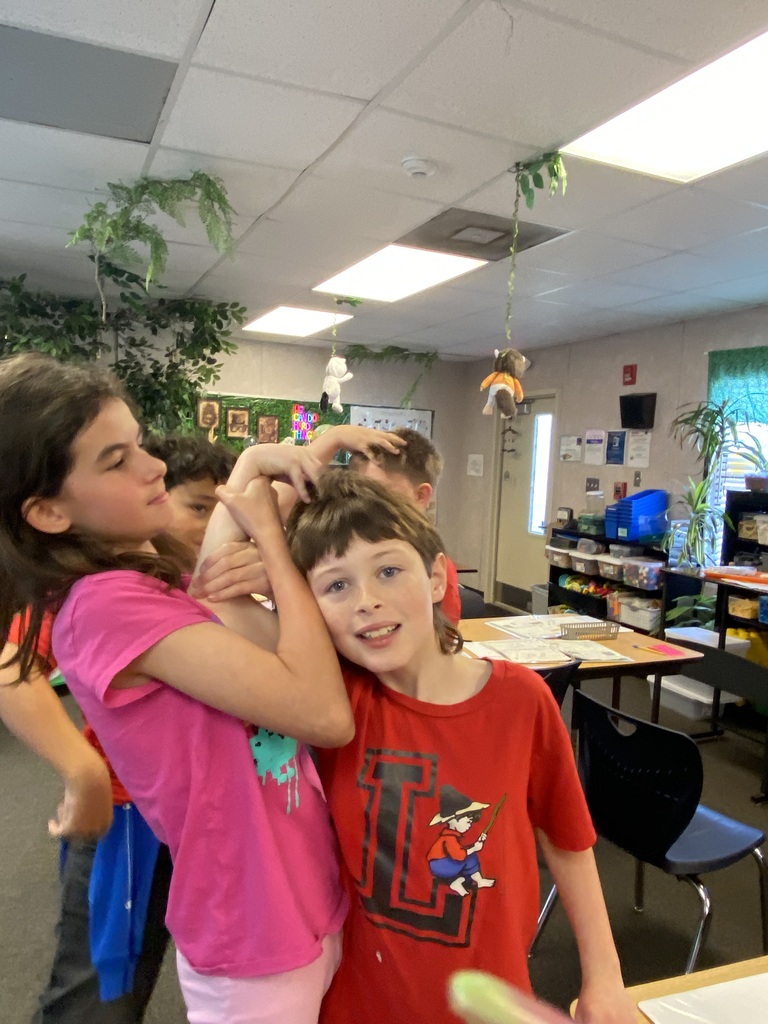 Two children, one with long hair, interact in a classroom. The girl has her hands on the boy's head.