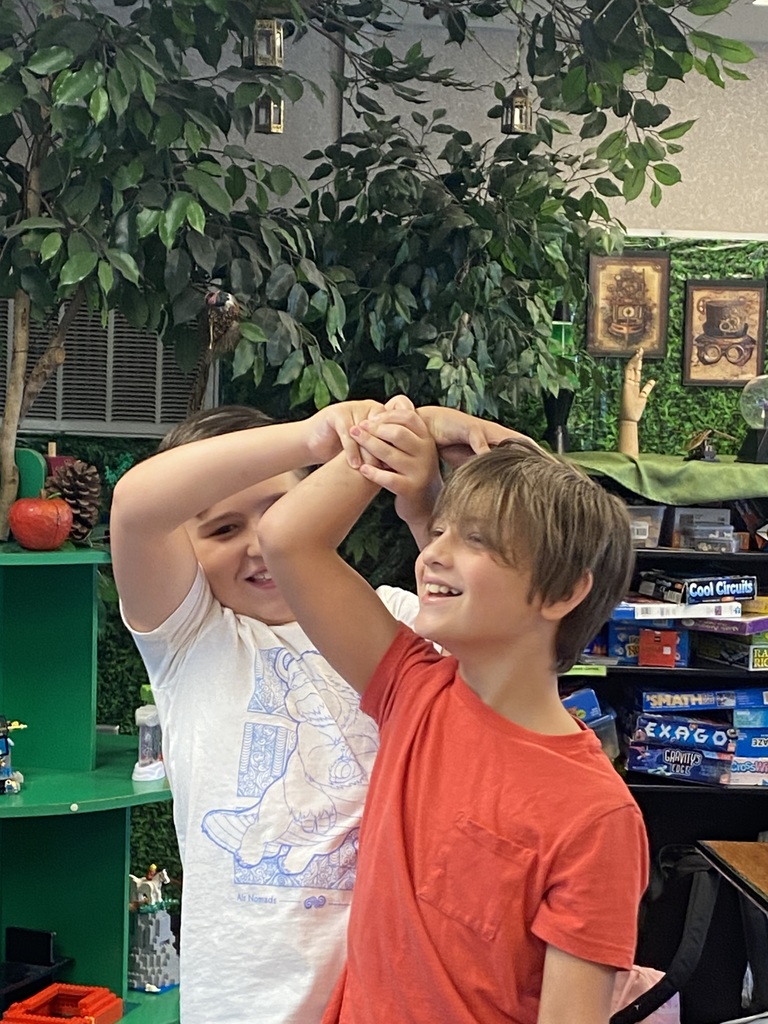 Two boys with hands clasped together, one in a white shirt, the other in orange. They are indoors.