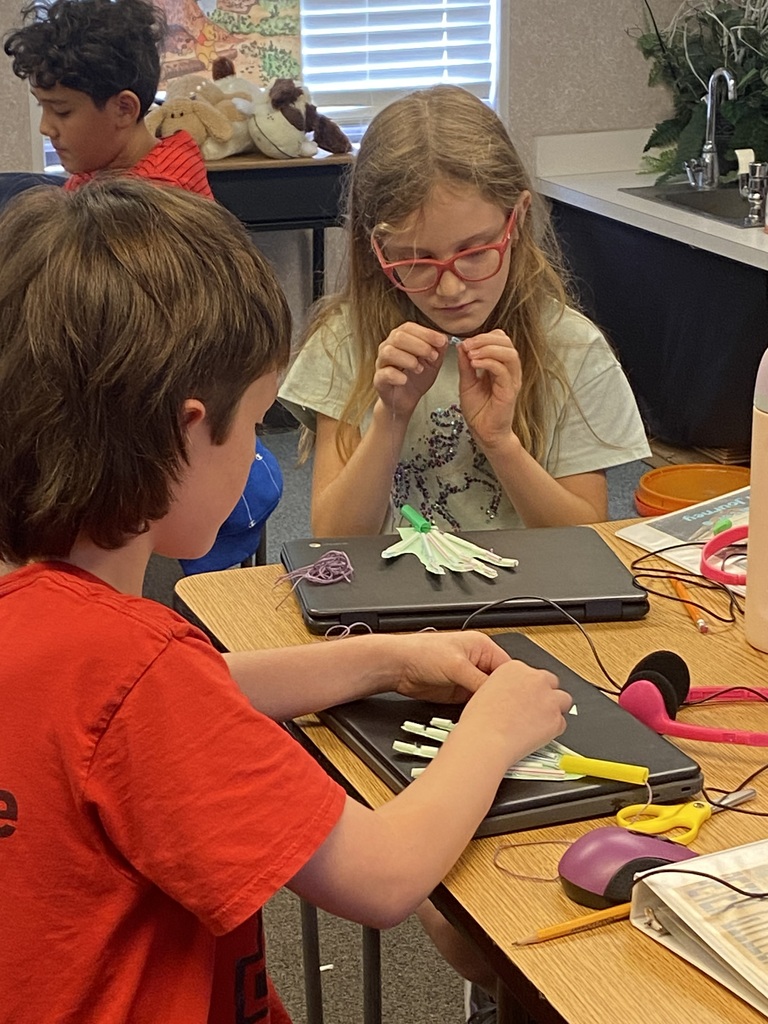 Kids in a classroom, one wearing glasses, are working on a laptop and a tablet.