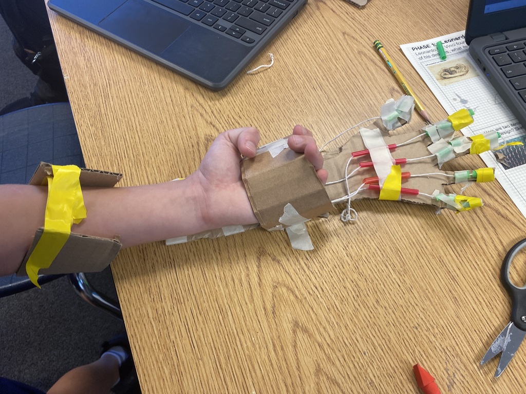 A person with a cardboard hand holding a tool and a laptop on a wooden table.