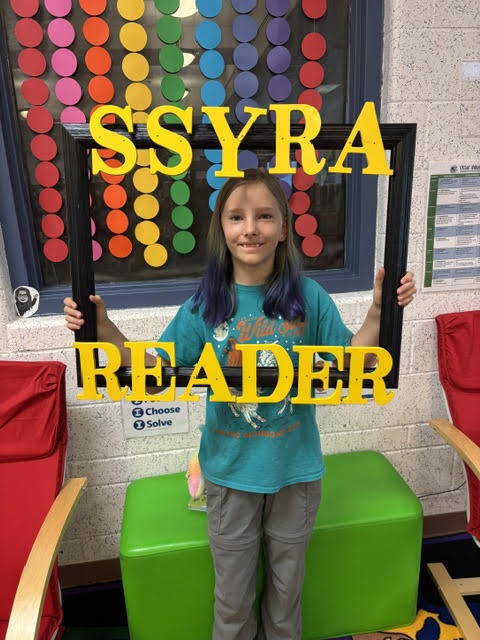 A girl holds a sign that reads "S.Syra Reader" in a classroom with a colorful circle decoration on the window.