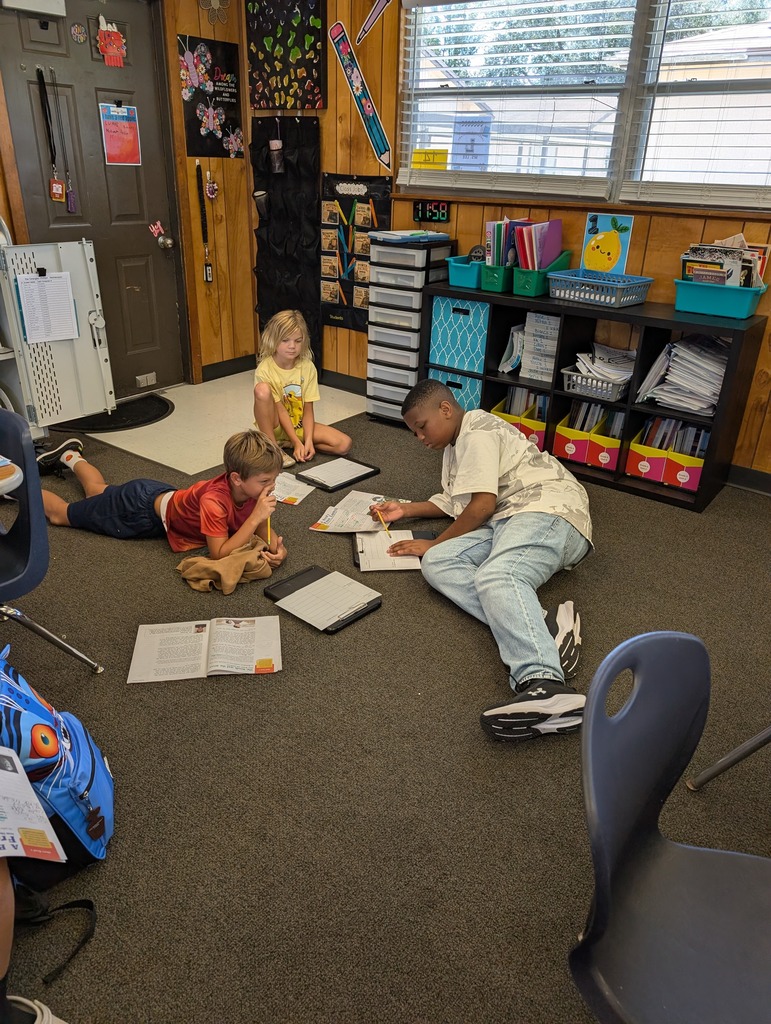 Four children on a classroom floor. One sitting and three lying down, all studying with papers.
