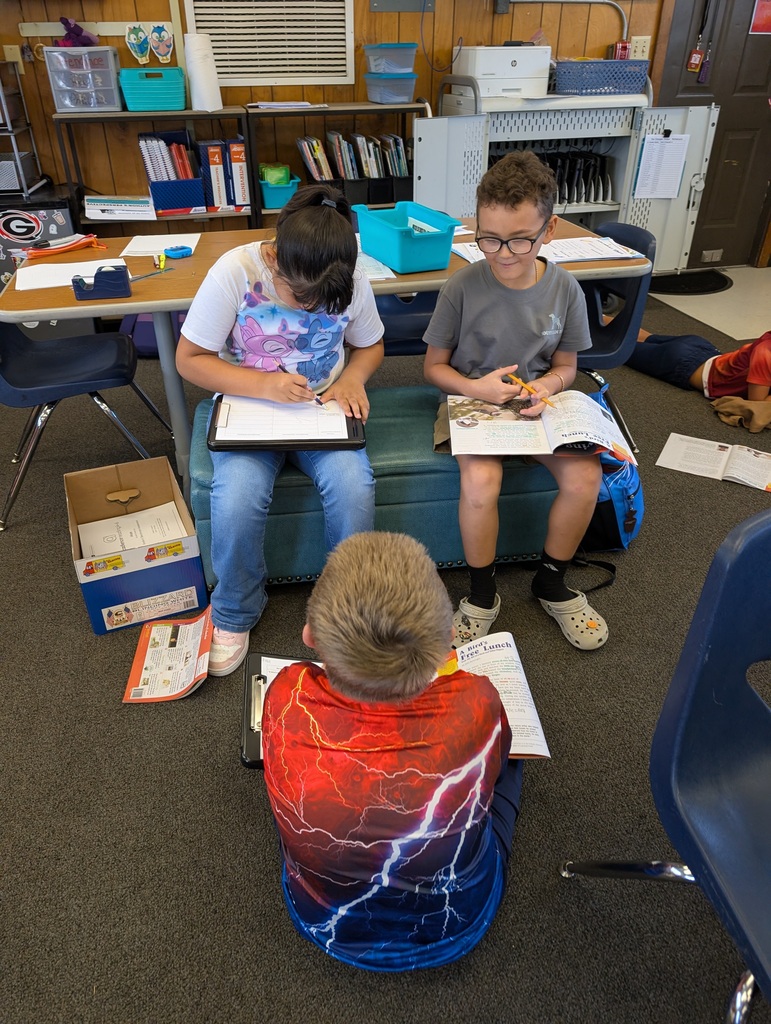 Three children sit on a bench in a classroom, each focused on their tasks, surrounded by shelves, tables, and chairs.