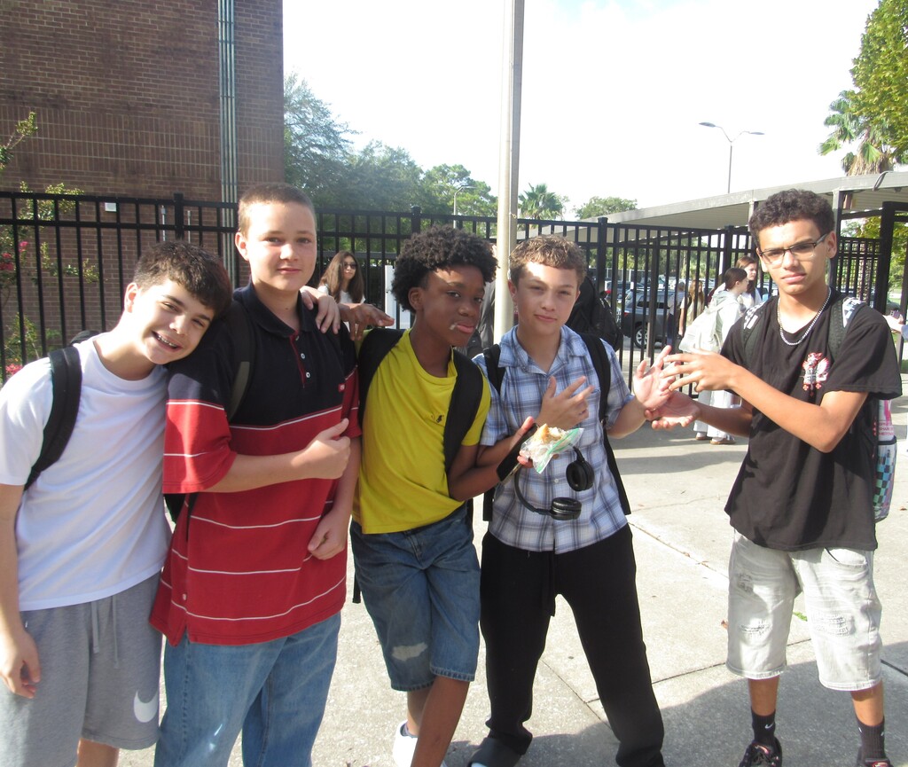 Five young males stand in front of a fence outside. One makes a peace sign.