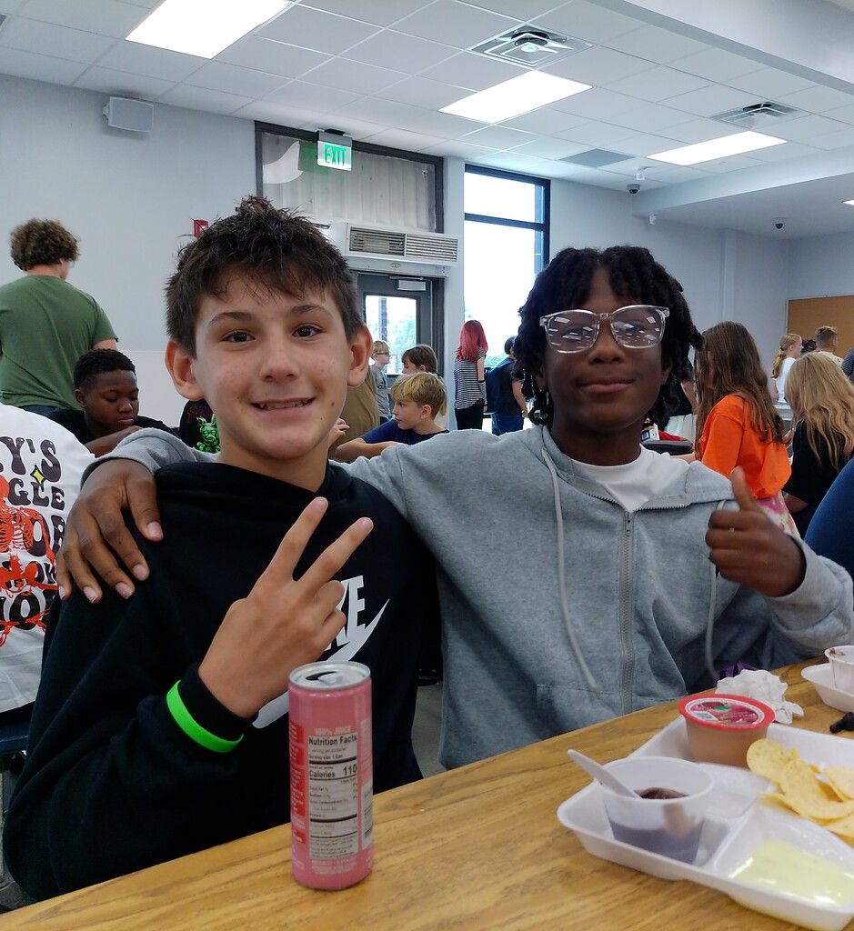 Two boys sitting at a cafeteria table, one making a peace sign, the other giving a thumbs up.