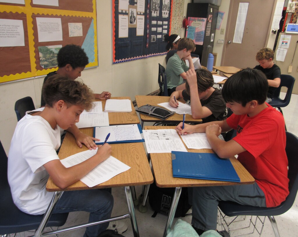 Classroom with desks. Students sit, writing on paper, with pens and books. Some look down, others at papers.