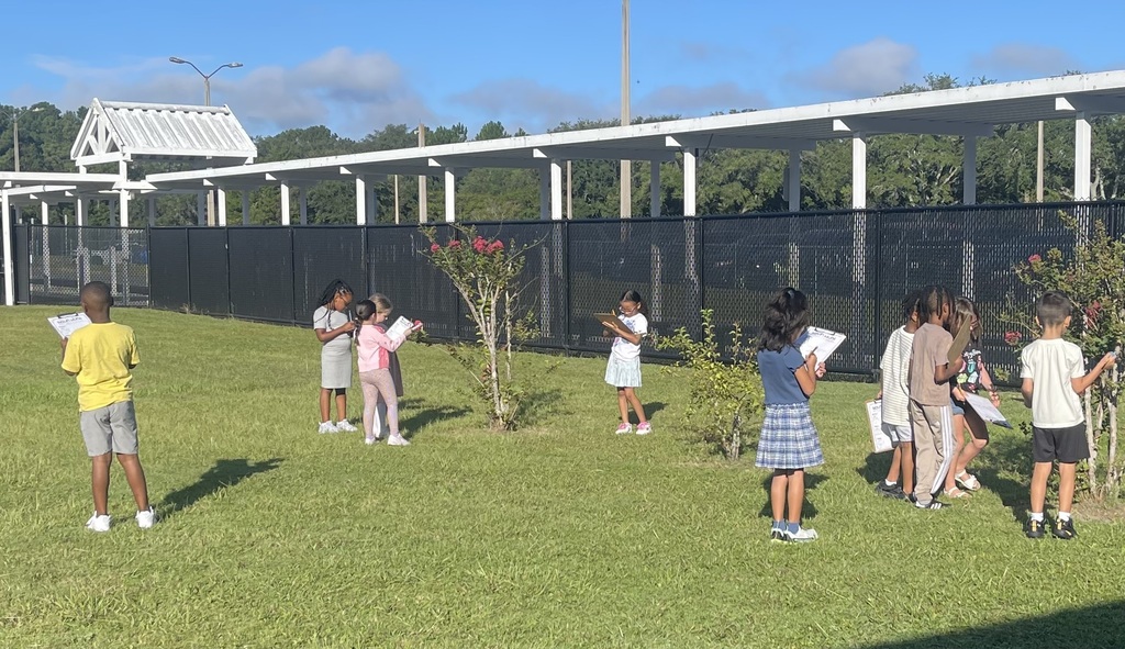 Group of children in a grassy area near a building and a fence with trees and shrubs.