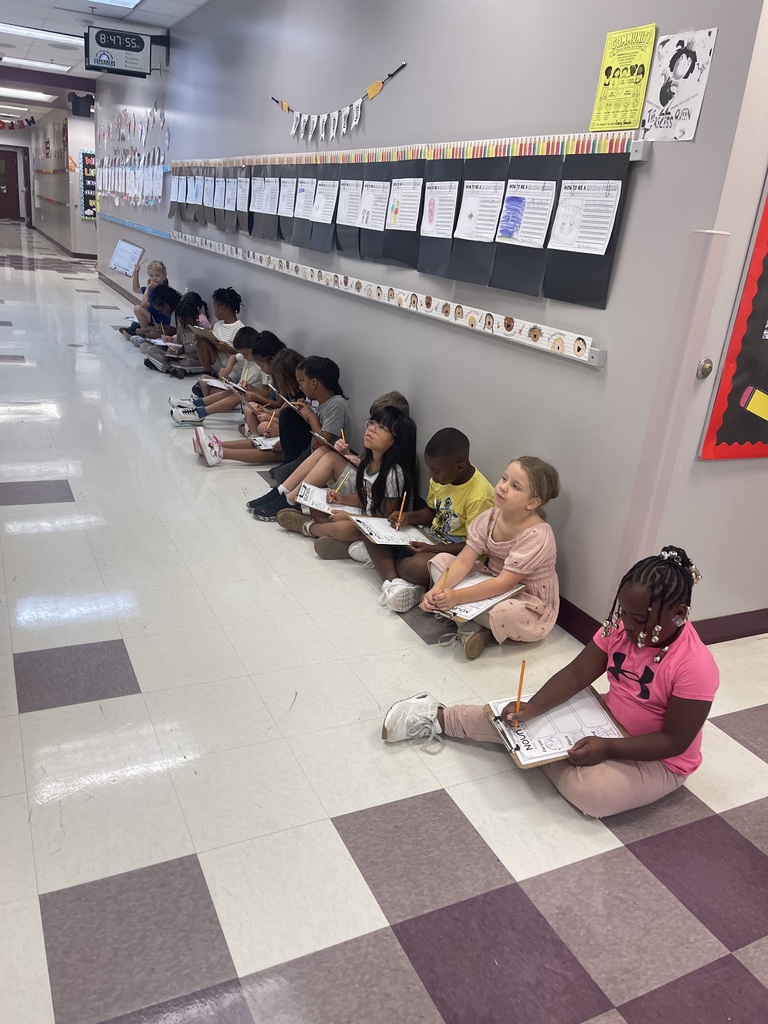 Students sit on a floor, facing a bulletin board with papers. They hold papers and pencils.