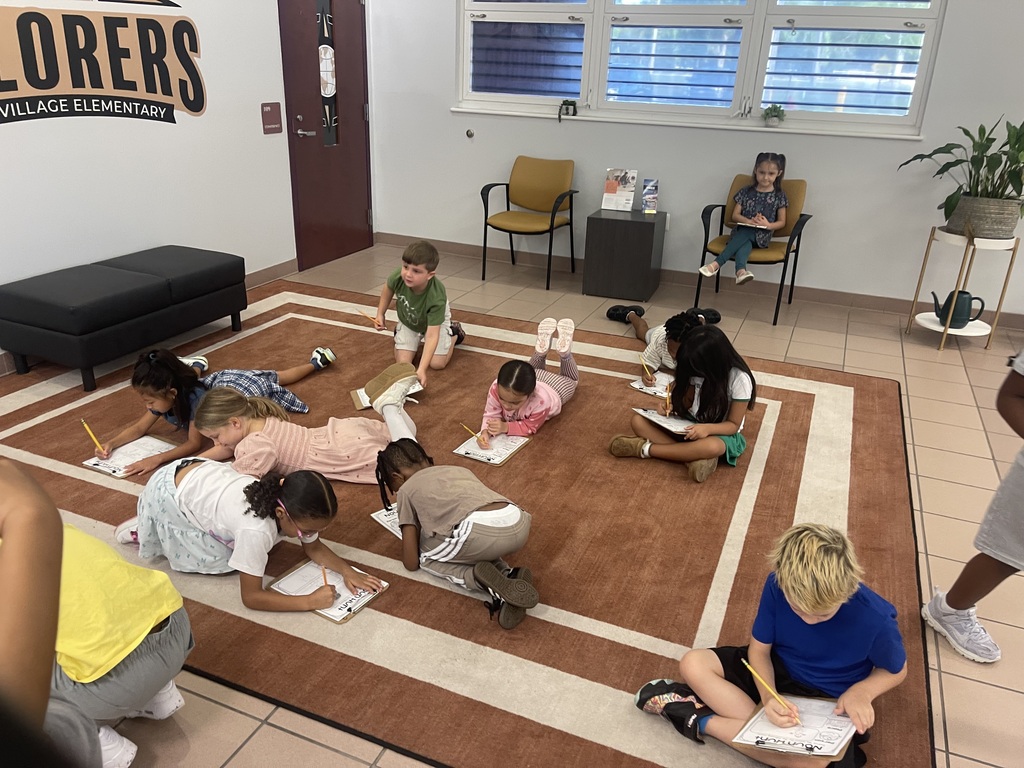 Group of children sitting on the floor and writing on paper, in a room with a rug and a window.