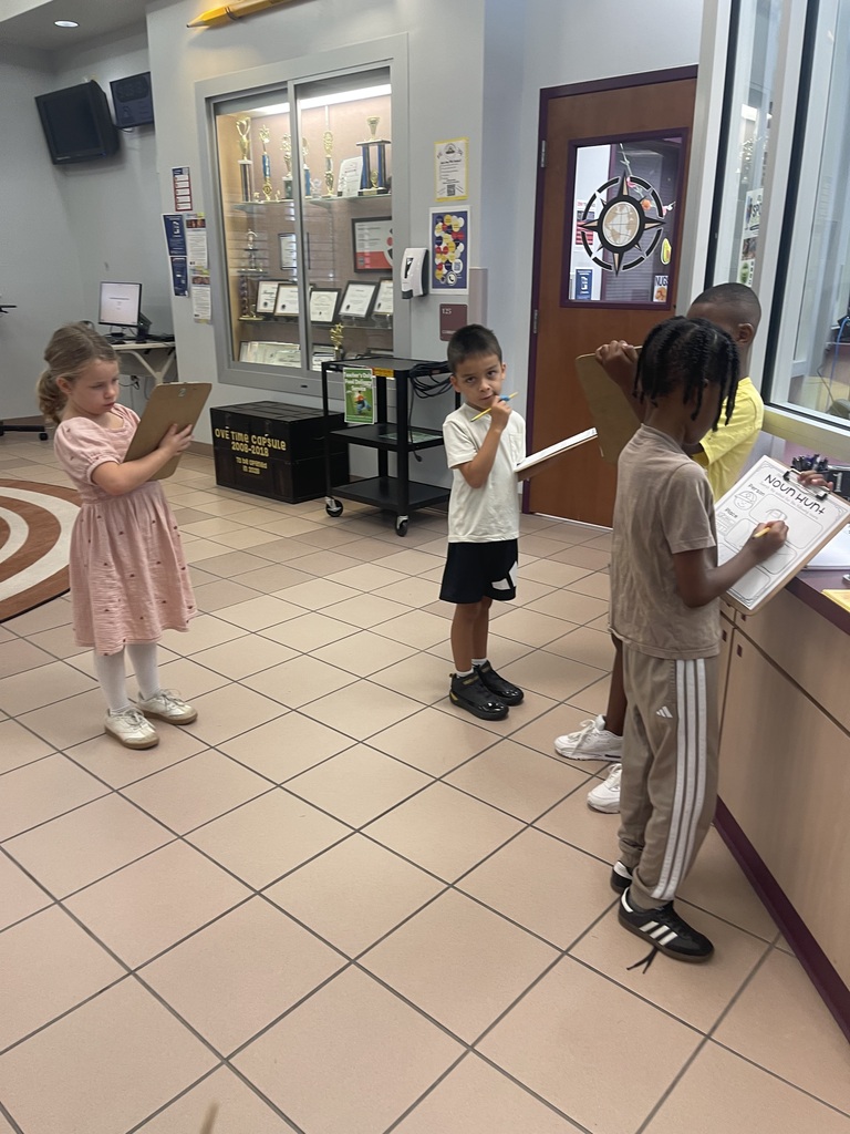 Four children stand in a room, holding clipboards. Two have their hands near their mouths. Tiled flooring and a door are visible.